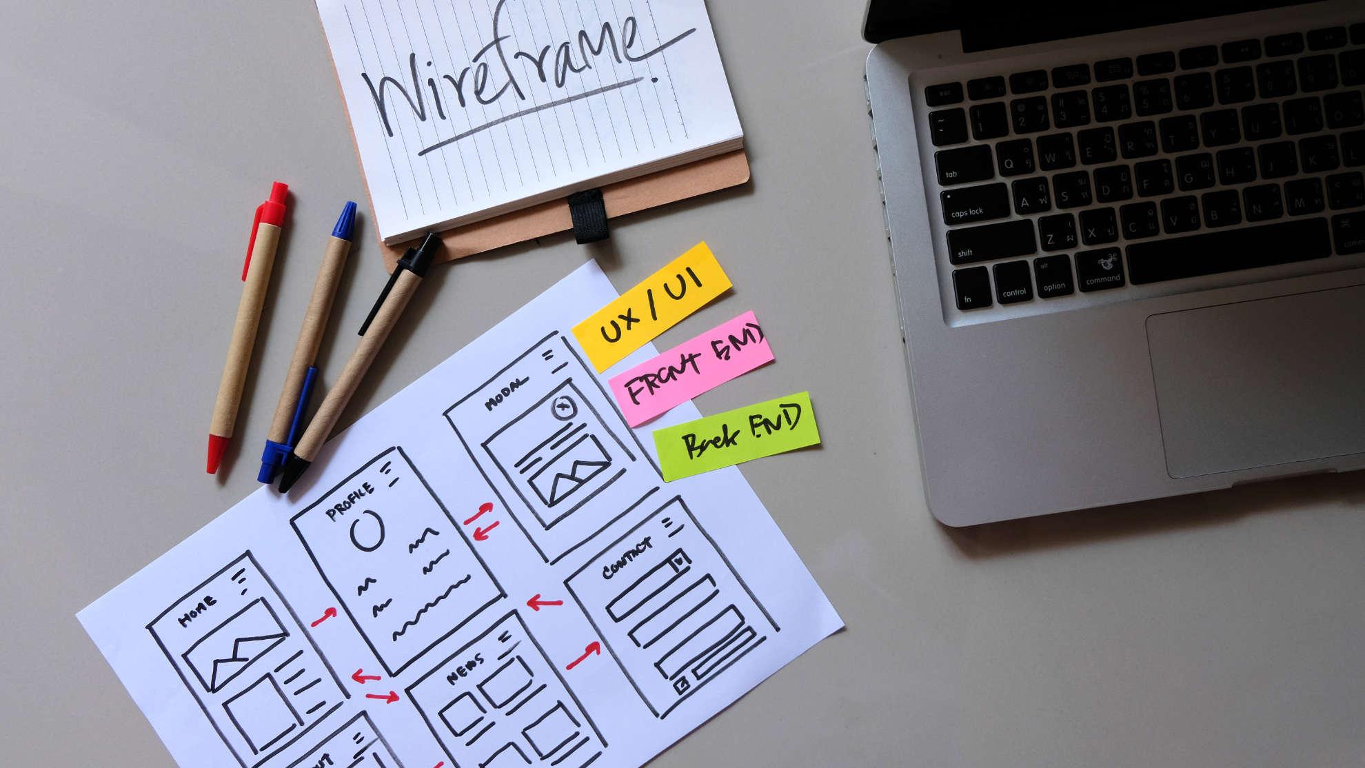 Hand-drawn website wireframes on paper next to pens and a laptop