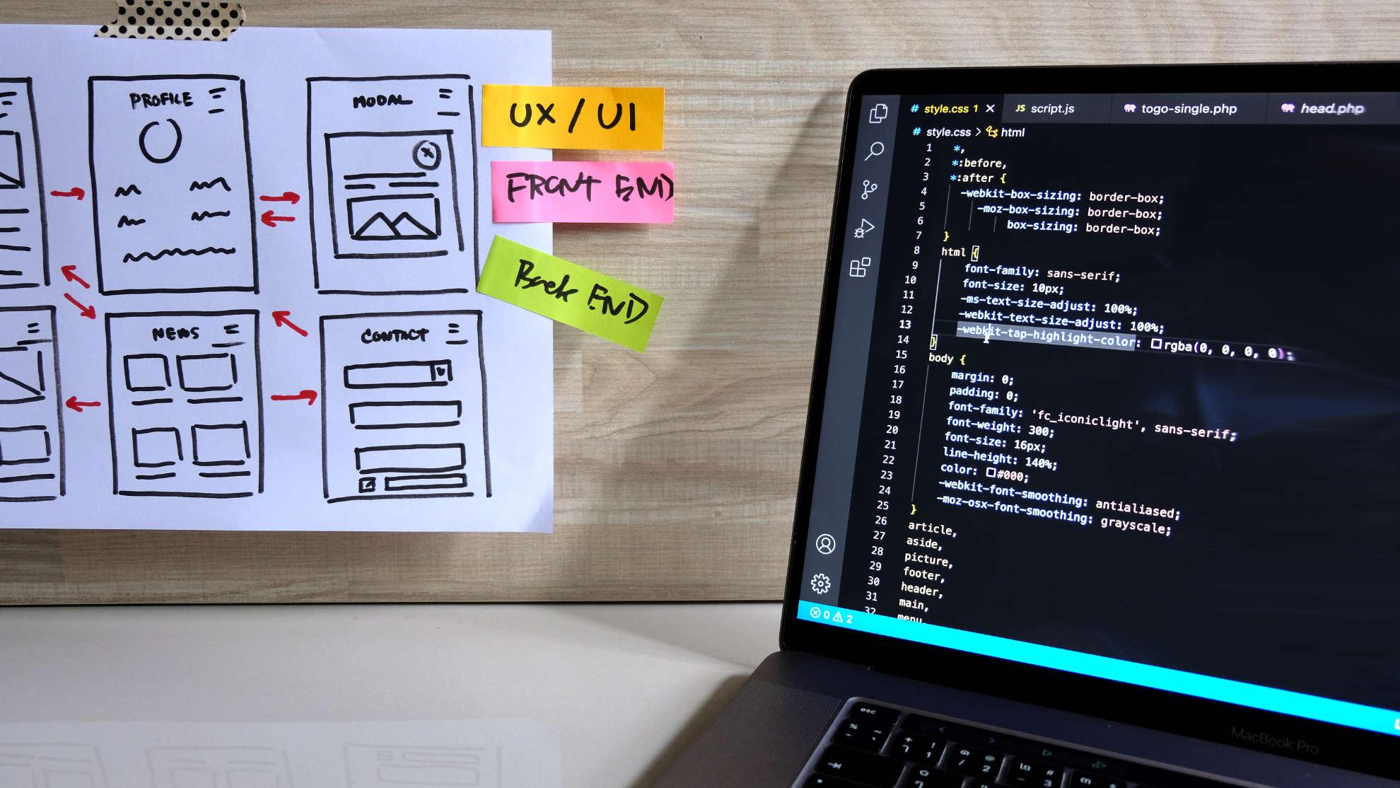Website wireframes and sticky notes next to a laptop showing code